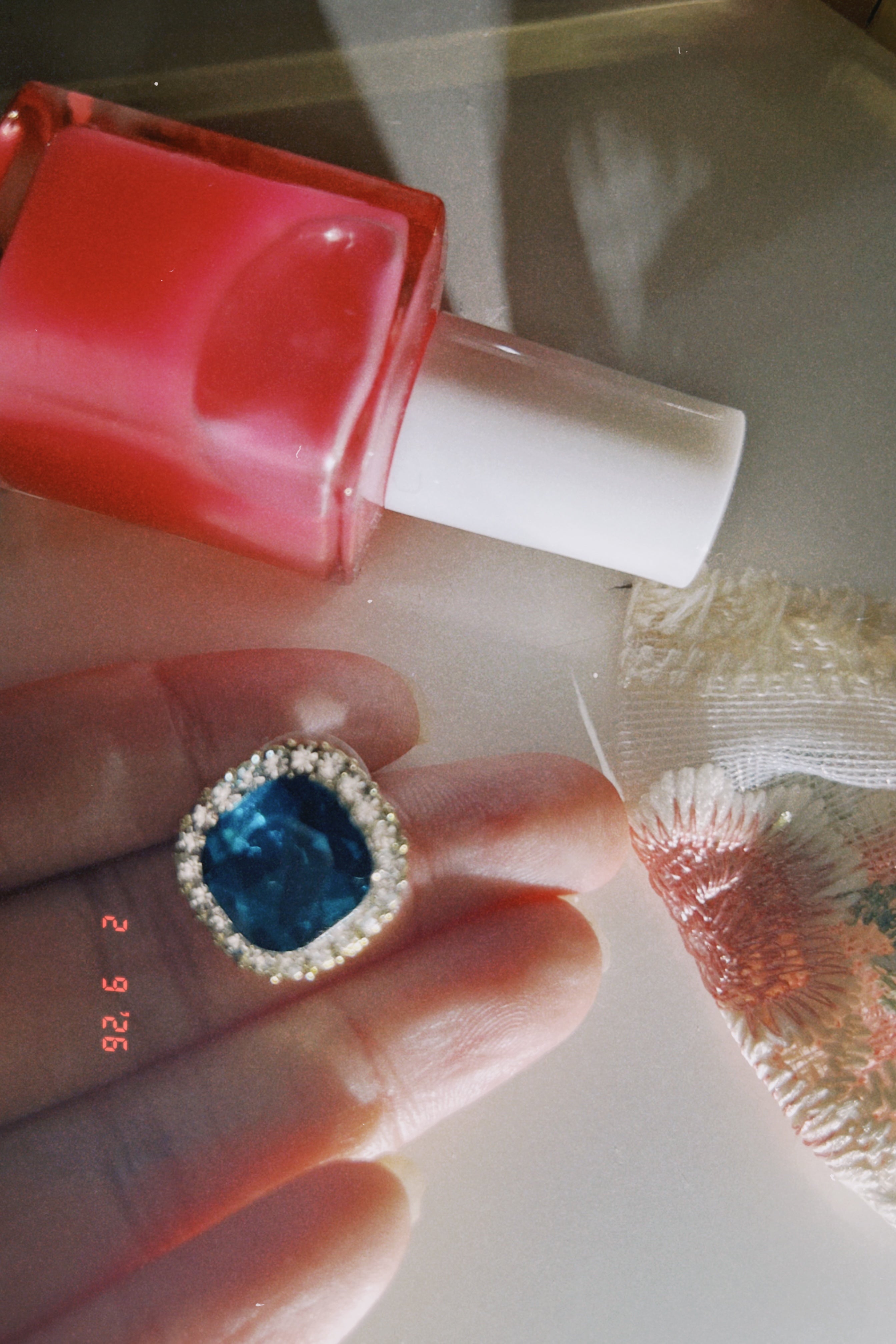 Close-up of a blue gemstone ring on a finger with a red nail polish bottle in the background.