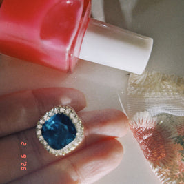 Close-up of a blue gemstone ring on a finger with a red nail polish bottle in the background.
