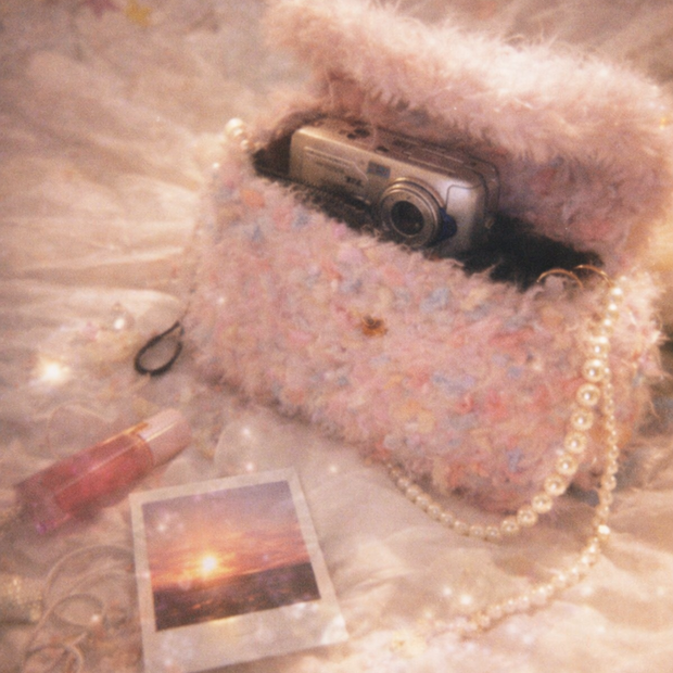 Someday she - Dream visual of the Deleted Scenes pink shaggy crochet bag on a soft dreamy surface, featuring a vintage digital camera and a sunset polaroid photo in an ethereal setting.