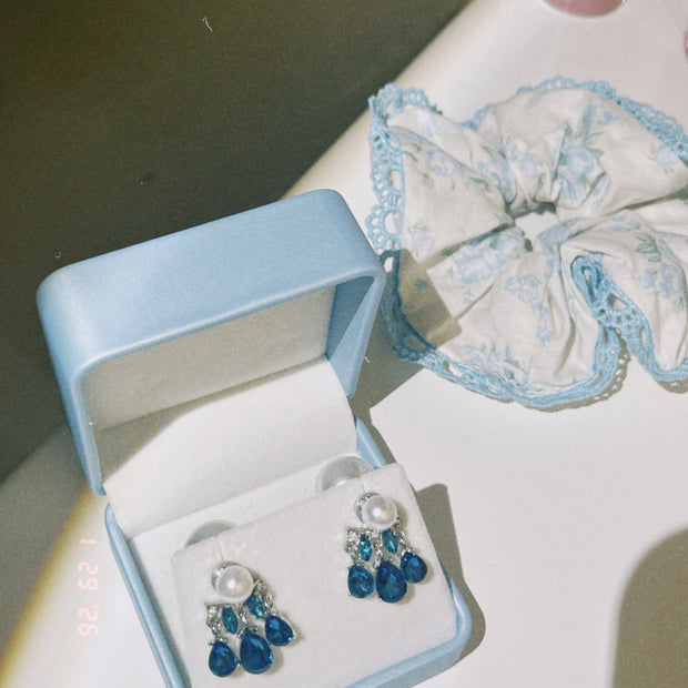 Someday she - Midnight Rain vintage-inspired silver chandelier earrings with deep blue gemstones and pearls, displayed in a light blue jewelry box next to a floral scrunchie.