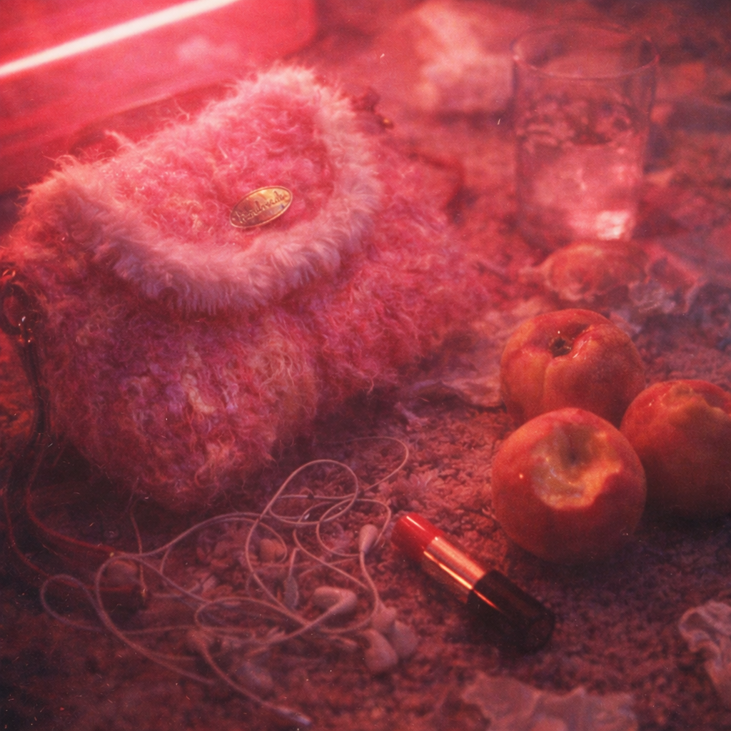 Someday she - Dream visual of the Peaches Never Sleep pink shaggy artisanal bag on a textured surface, surrounded by bitten red apples, a lipstick, and tangled earphones in a dramatic deep red light.