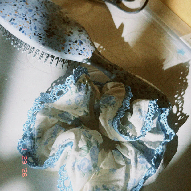 Someday she - The Alice coquette aesthetic blue floral scrunchie with delicate lace ruffled trimming, resting on a sunlit surface next to a blue patterned hairbrush with soft shadows.
