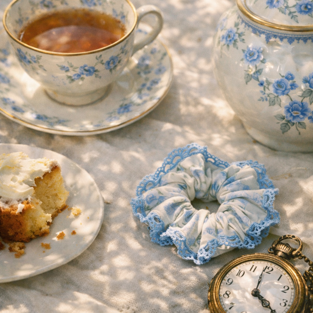 Someday she - Dream visual of The Alice blue floral lace-trimmed ruffled hair tie at a vintage picnic with a floral teacup, teapot, a slice of cake, and a golden pocket watch on a white tablecloth in the grass.