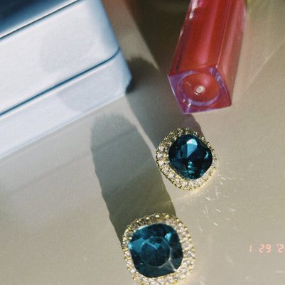 Someday she - The Blue Hour vintage-style square stud earrings featuring deep blue gemstones with a sparkling gold rim, resting on a reflective surface next to a light blue jewelry box.