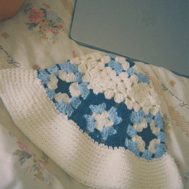 Someday she - The Cloud Watcher hand-crocheted granny square bucket hat with a blue and white floral pattern, resting on a soft floral bedspread next to a laptop.