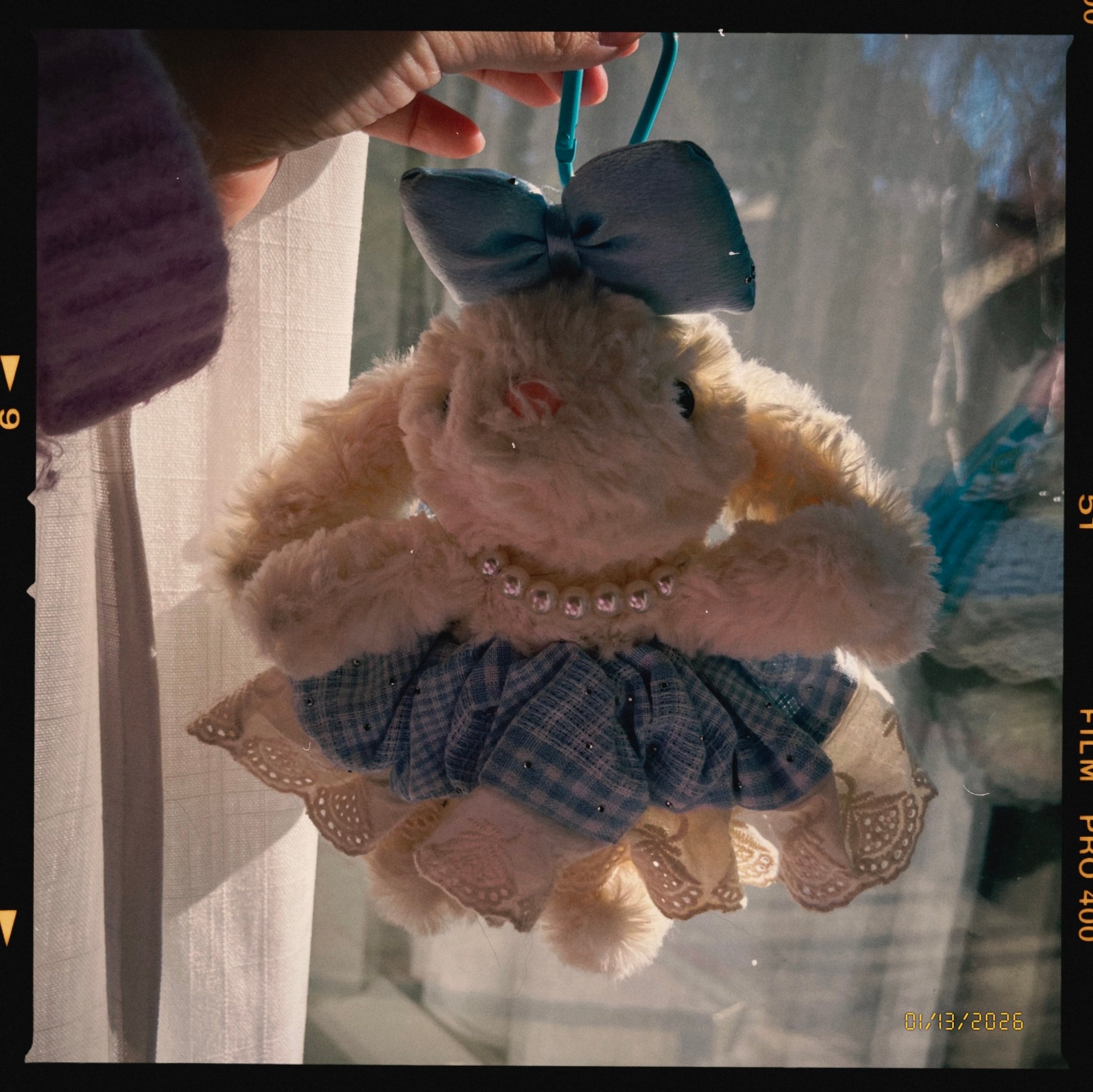Someday she - The Imaginary Friend vintage fluffy bunny bag charm with a blue bow and plaid dress, held by a hand against soft window light.
