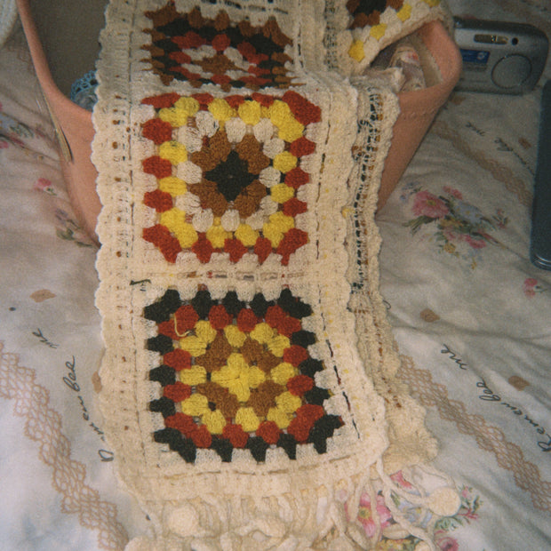 Someday she - The Little Women artisanal hand-crocheted scarf featuring colorful geometric granny squares in autumn tones, draped over a soft storage bin in a warm setting.