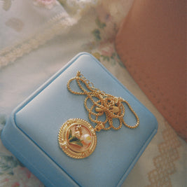 Someday she - The Love Letter French vintage relief floral medallion necklace in gold, resting on a light blue jewelry box with a delicate floral lace and pearl backdrop.